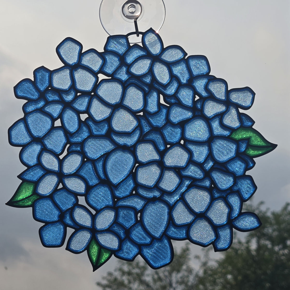 A 3D printed suncatcher shaped like a hydrangea, featuring shades of blue and light blue petals with green leaves, suspended by a clear suction cup against an outdoor sky background.