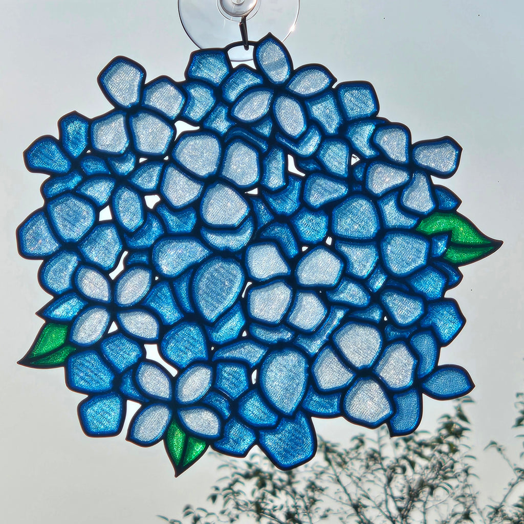 A 3D printed suncatcher shaped like a hydrangea, featuring shades of blue, light blue, and green leaves, suspended from a clear suction cup against an outdoor sky background.