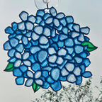 A 3D printed suncatcher shaped like a hydrangea, featuring shades of blue, light blue, and green leaves, suspended from a clear suction cup against an outdoor sky background.
