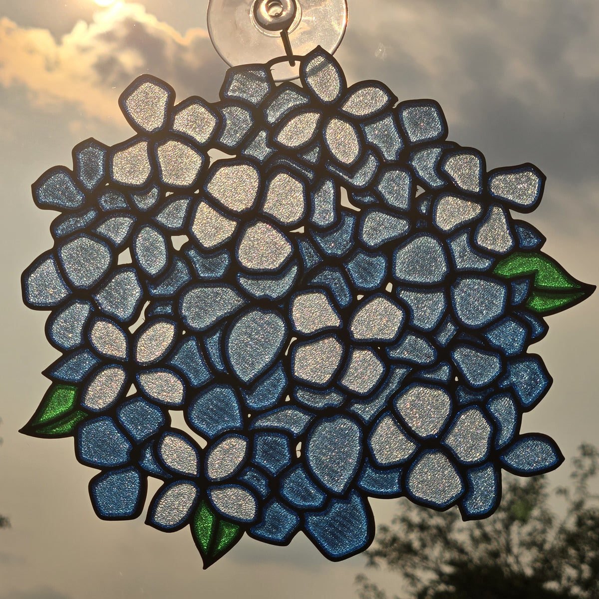 A 3D printed suncatcher shaped like a hydrangea, featuring blue and white petals with green leaves and dark outlines, suspended by a clear suction cup against a cloudy sky.