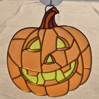 A 3D printed pumpkin ornament with a carved jack-o'-lantern face, featuring orange and brown glass panels with light green glowing eyes and mouth details, hanging against a neutral fabric background.