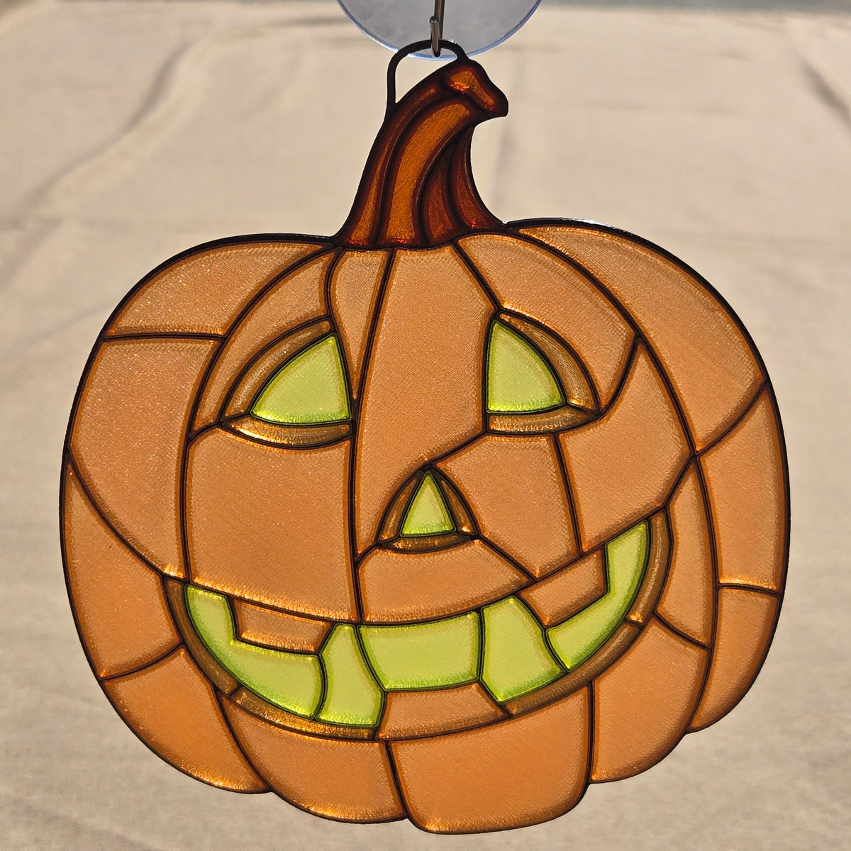A 3D printed pumpkin ornament with a carved jack-o'-lantern face, featuring orange and brown glass panels with light green glowing eyes and mouth details, hanging against a neutral fabric background.
