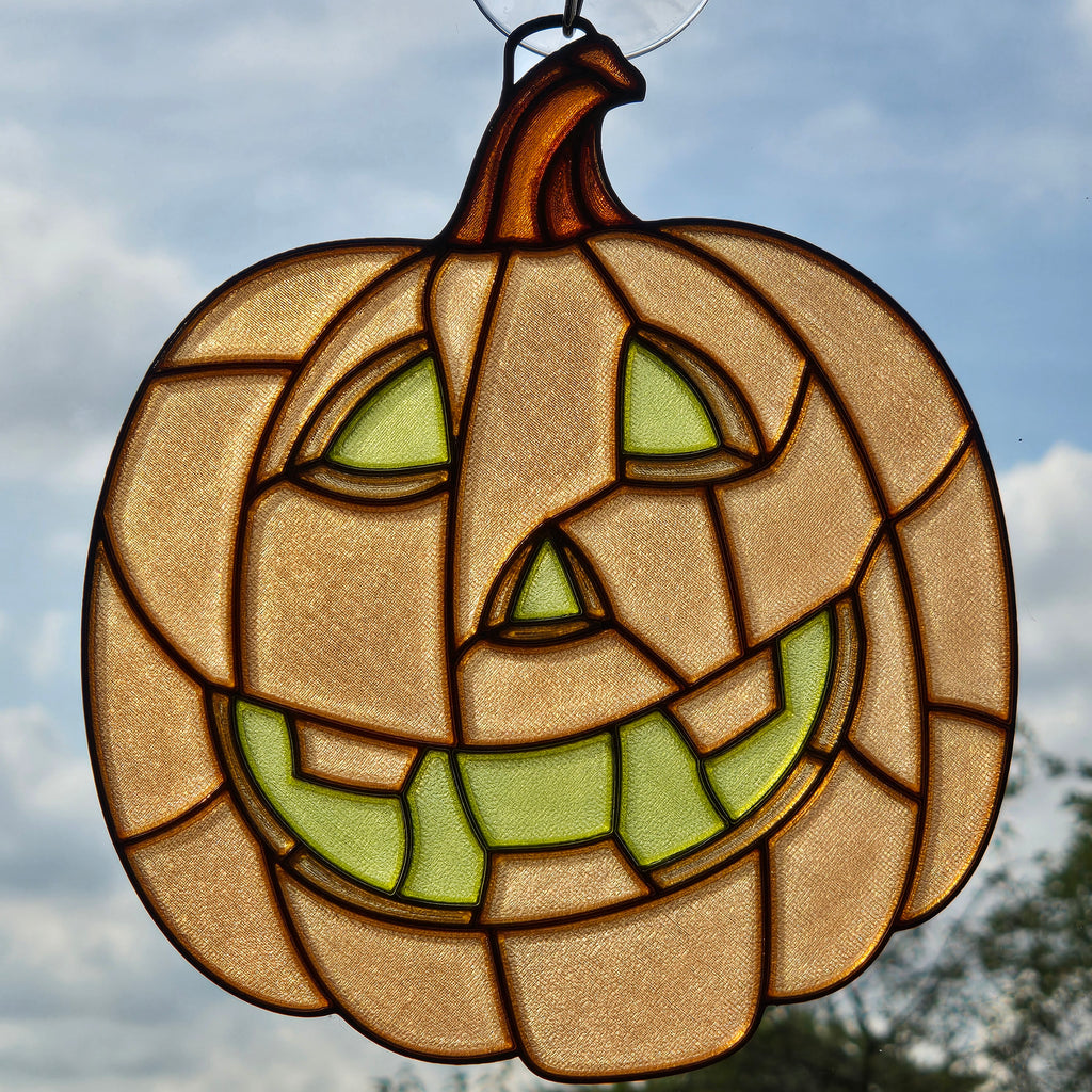 A 3D printed pumpkin hanging ornament with a carved jack-o'-lantern face, featuring a warm tan textured glass body, green glass eyes and mouth, and dark brown lead lines, shown against an outdoor cloudy sky background.