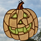 A 3D printed pumpkin hanging ornament with a carved jack-o'-lantern face, featuring a warm tan textured glass body, green glass eyes and mouth, and dark brown lead lines, shown against an outdoor cloudy sky background.