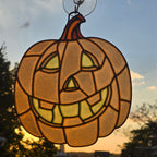 A 3D printed pumpkin suncatcher with a carved jack-o'-lantern face, hanging from a clear suction cup. The glass panels are warm amber and yellow, backlit by a sunset sky with blue and orange tones.