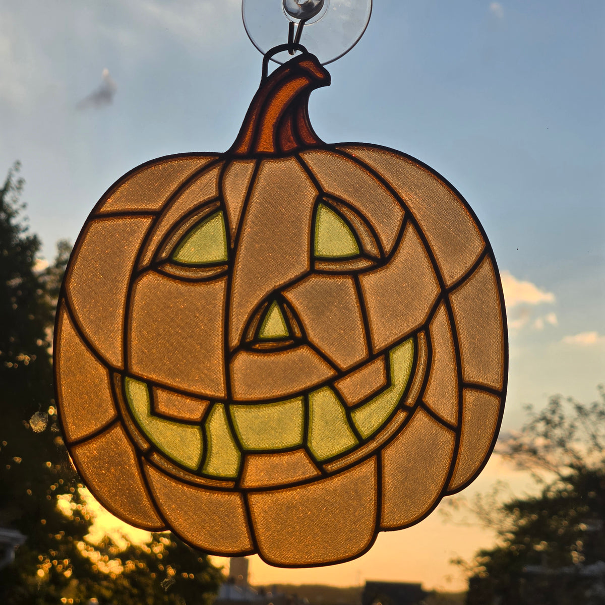 A 3D printed pumpkin suncatcher with a carved jack-o'-lantern face, hanging from a clear suction cup. The glass panels are warm amber and yellow, backlit by a sunset sky with blue and orange tones.