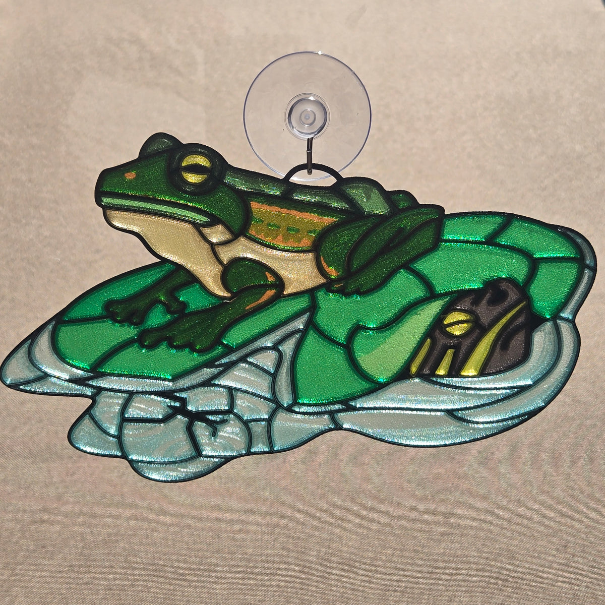 A 3d printed suncatcher featuring a green tree frog sitting on a green leaf, attached to a clear suction cup at the top, set against a neutral tan background.