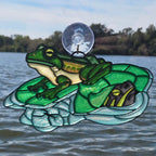 A 3d printed window ornament depicting a green frog sitting on a turtle shell, attached to a clear suction cup and displayed against a window overlooking a lake.