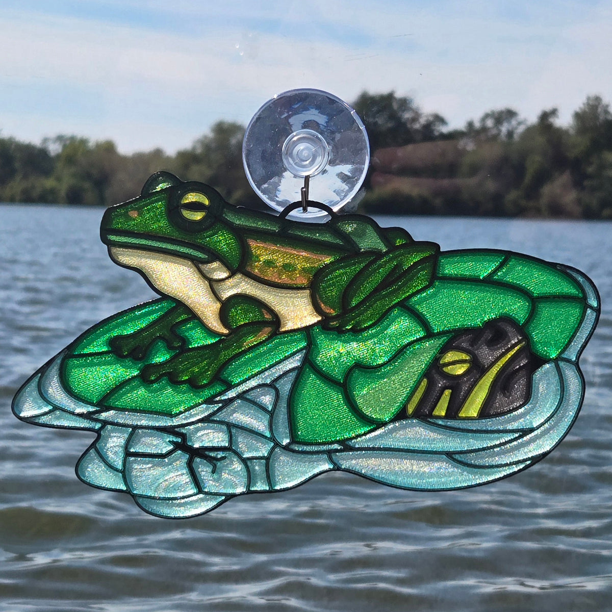 A 3d printed window ornament depicting a green frog sitting on a turtle shell, attached to a clear suction cup and displayed against a window overlooking a lake.