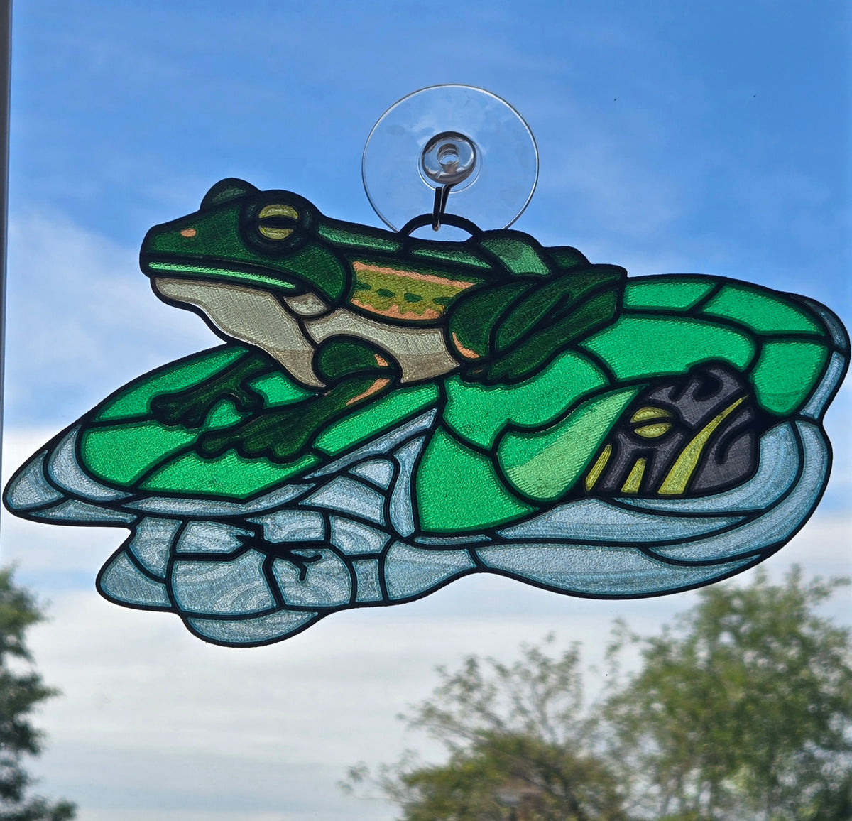 A 3d printed suncatcher featuring a green tree frog sitting on a winged insect, suspended from a clear suction cup against a blue sky background.