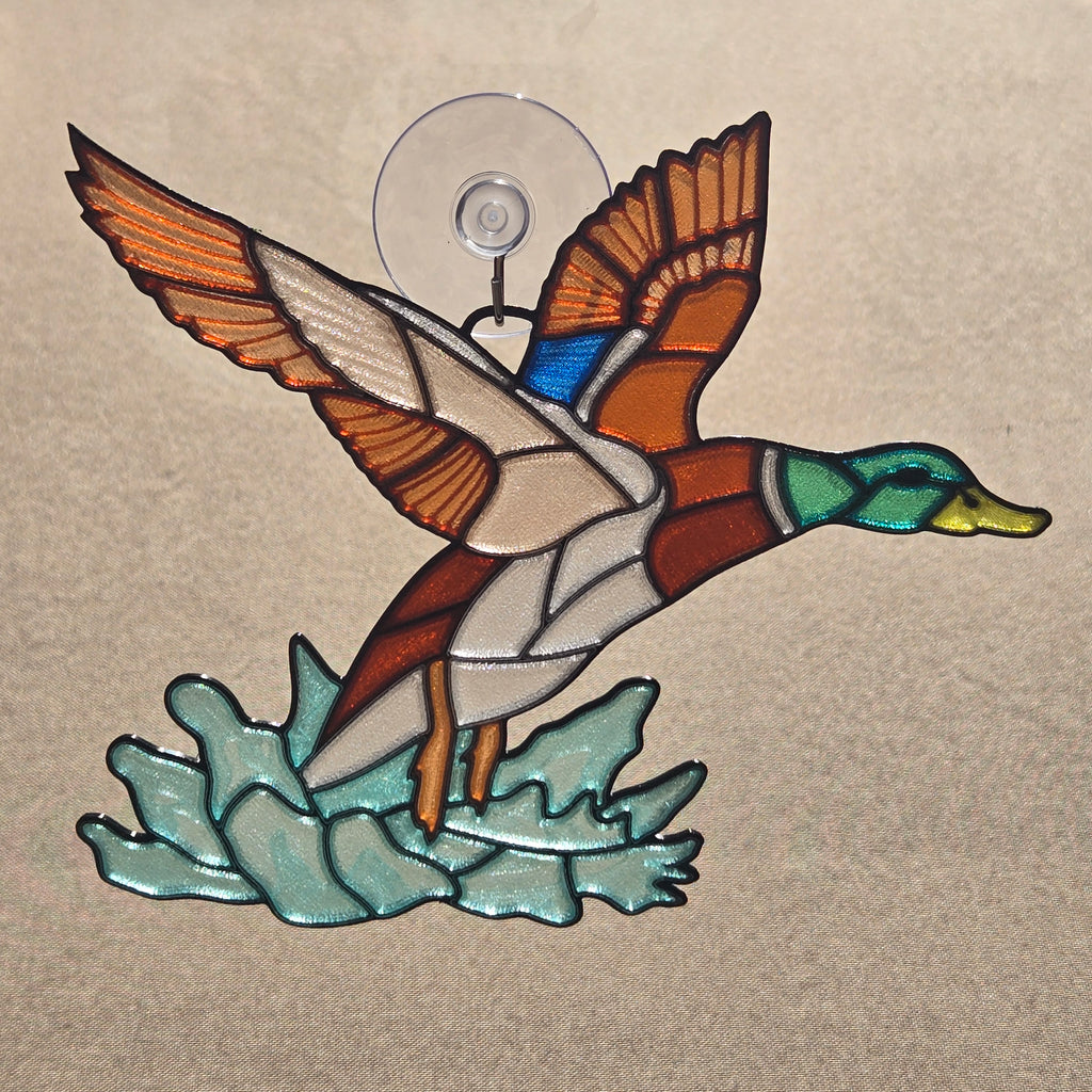 A 3D printed window piece depicting a mallard duck in flight, mounted with a clear suction cup on a beige textured background.