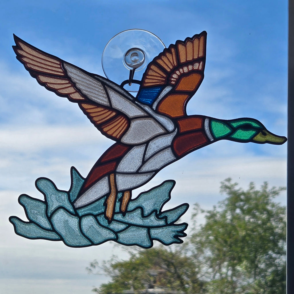A 3d printed window panel depicting a flying mallard duck in flight over blue water, mounted with a clear suction cup against an outdoor sky background.