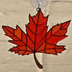 A 3D printed maple leaf ornament in shades of red and orange, suspended from a clear suction cup against a beige fabric background.