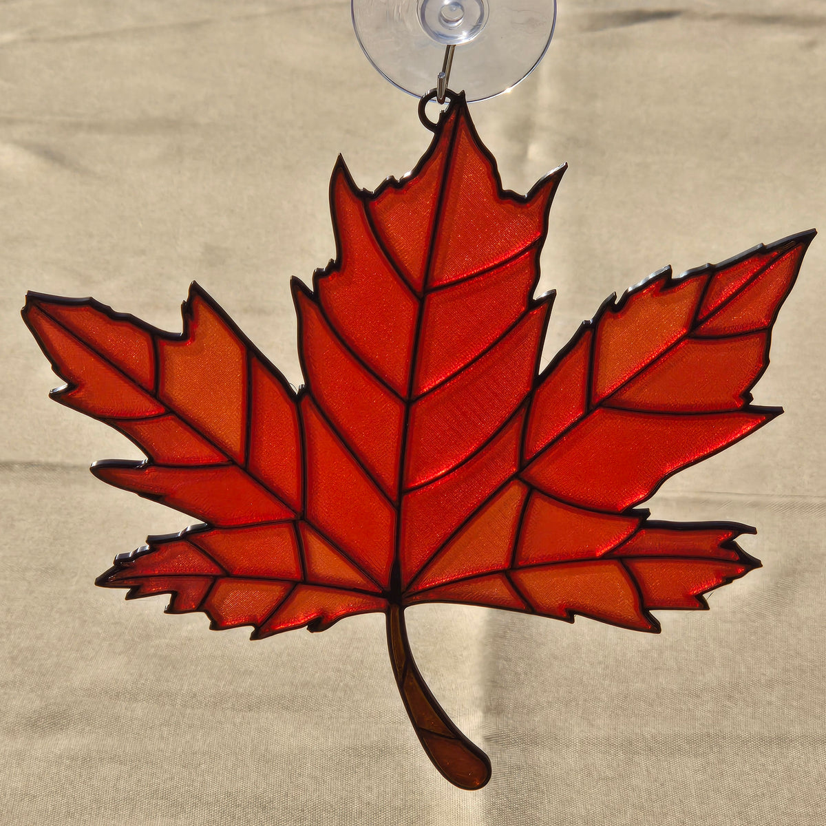 A 3D printed maple leaf ornament in shades of red and orange, suspended from a clear suction cup against a beige fabric background.