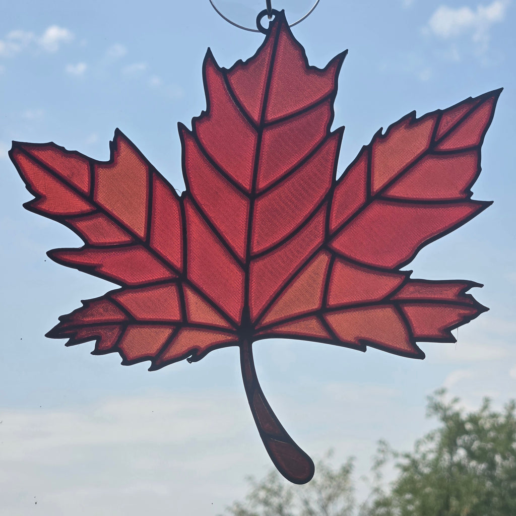 A red maple leaf suncatcher with black outlines and a hanging loop at the top, shown against a blue sky background.
