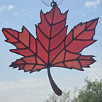 A red maple leaf suncatcher with black outlines and a hanging loop at the top, shown against a blue sky background.