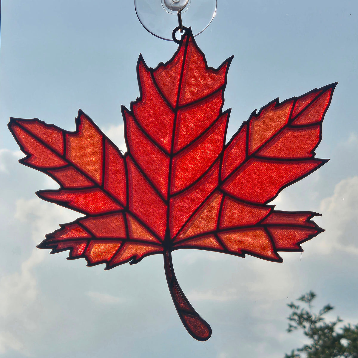 A red maple leaf 3D printed suncatcher hanging ornament suspended by a clear glass loop against a cloudy sky background.