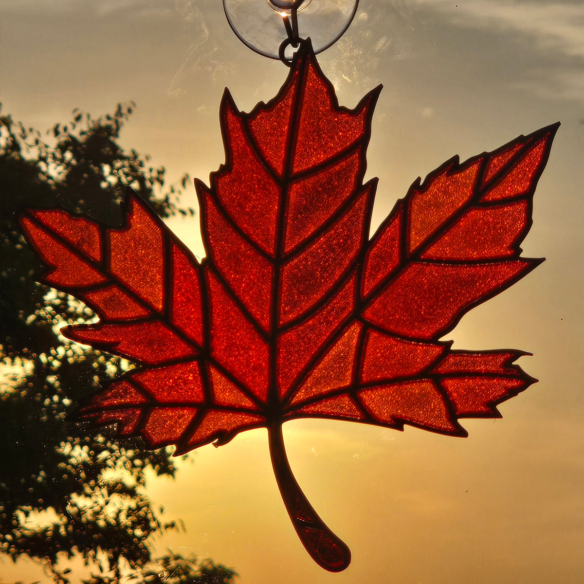 A red glitter maple leaf suncatcher hanging in front of a warm sunset sky with tree silhouettes.