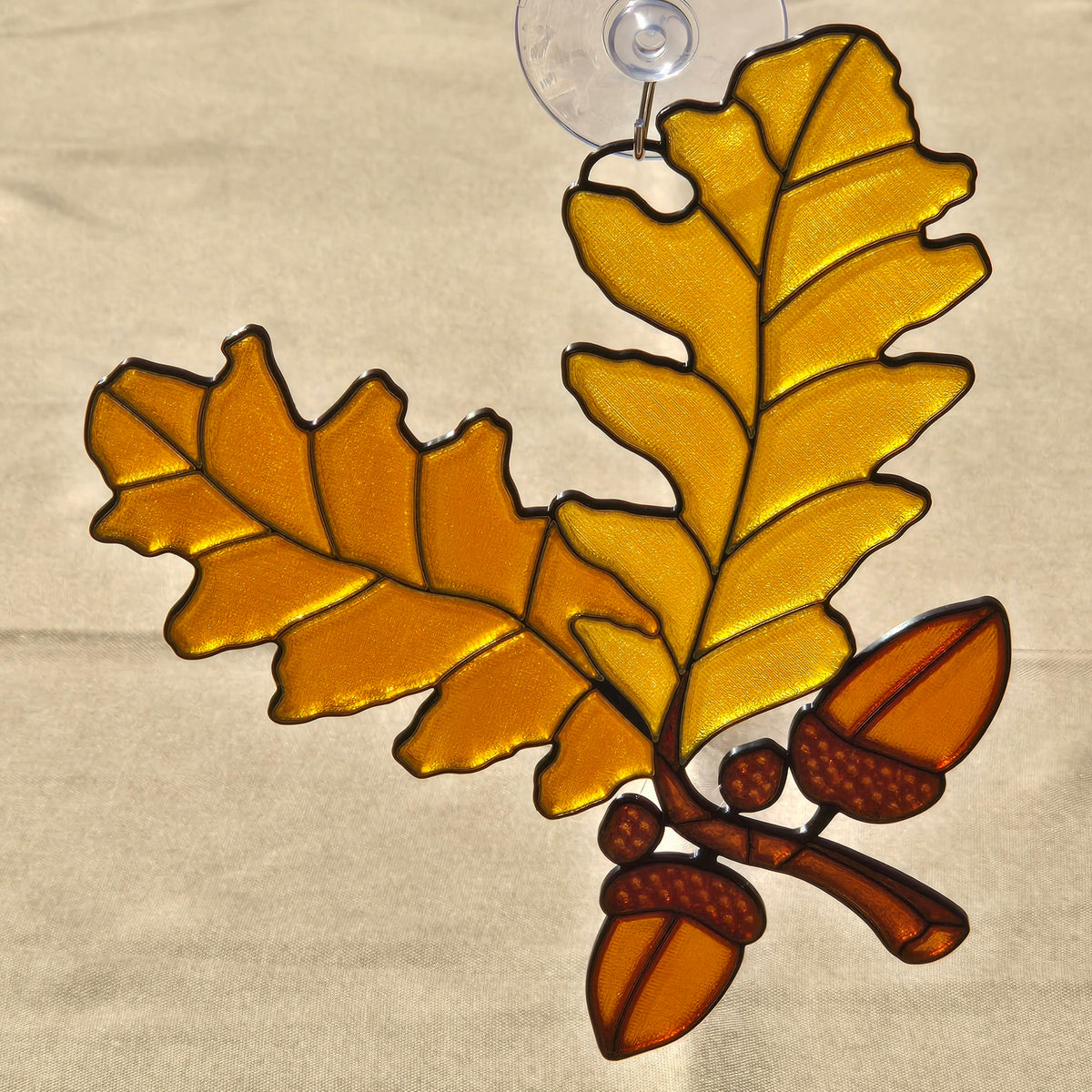 A 3D printed ornament depicting an oak branch with two large golden-yellow leaves and three brown acorns, outlined in dark metal and suspended from a clear suction cup.