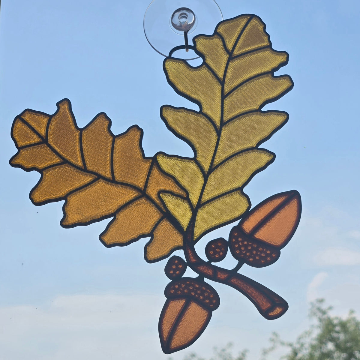 A 3D printed window ornament depicting an oak branch with two golden-yellow leaves, brown acorns, and dark outlines, suspended by a clear suction cup against a blue sky background.