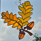 A 3D printed window ornament depicting two golden oak leaves and two acorns on a branch, suspended by a clear glass loop against a blue sky with clouds.