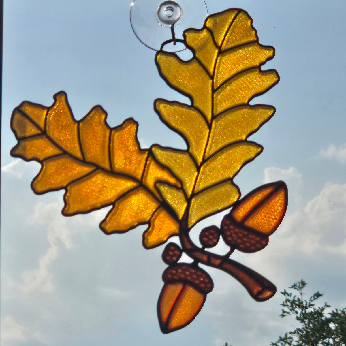 A 3D printed window ornament depicting two golden oak leaves and two acorns on a branch, suspended by a clear glass loop against a blue sky with clouds.