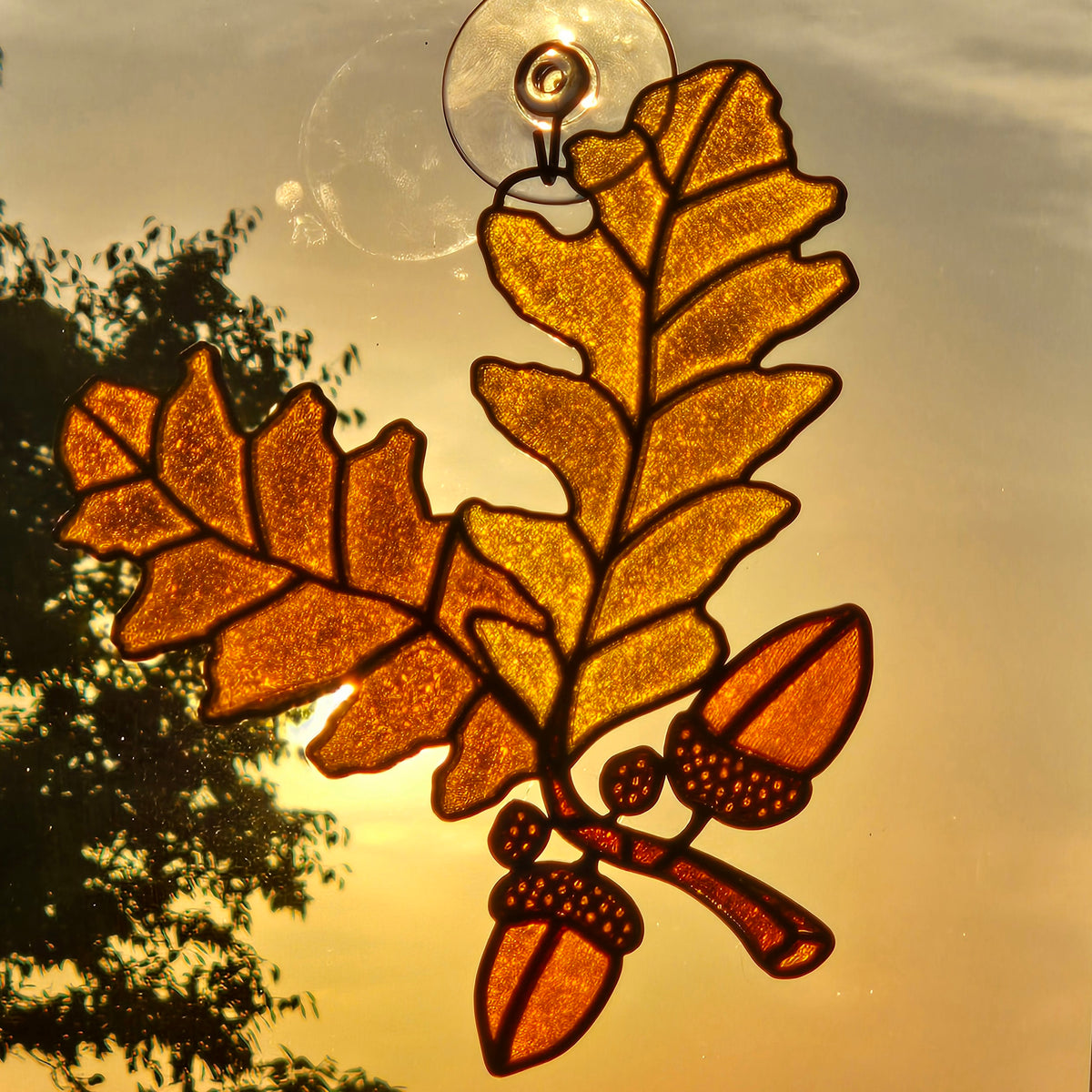 An autumn-themed 3D printed ornament featuring oak leaves and acorns, suspended from a clear suction cup against a warm sunset sky with tree silhouettes.
