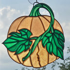 A 3D printed pumpkin suncatcher with a green stem, leaves, and a kissing face motif, shown hanging against a cloudy sky.