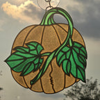 A pumpkin-shaped 3D printed suncatcher hanging ornament with green leaves and stem, suspended by a clear hook against an outdoor cloudy sky background.
