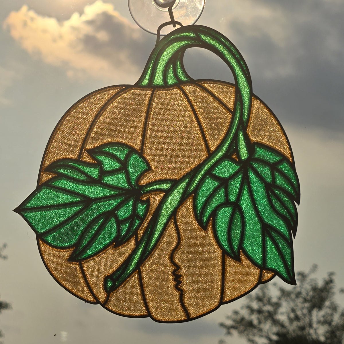 A pumpkin-shaped 3D printed suncatcher hanging ornament with green leaves and stem, suspended by a clear hook against an outdoor cloudy sky background.