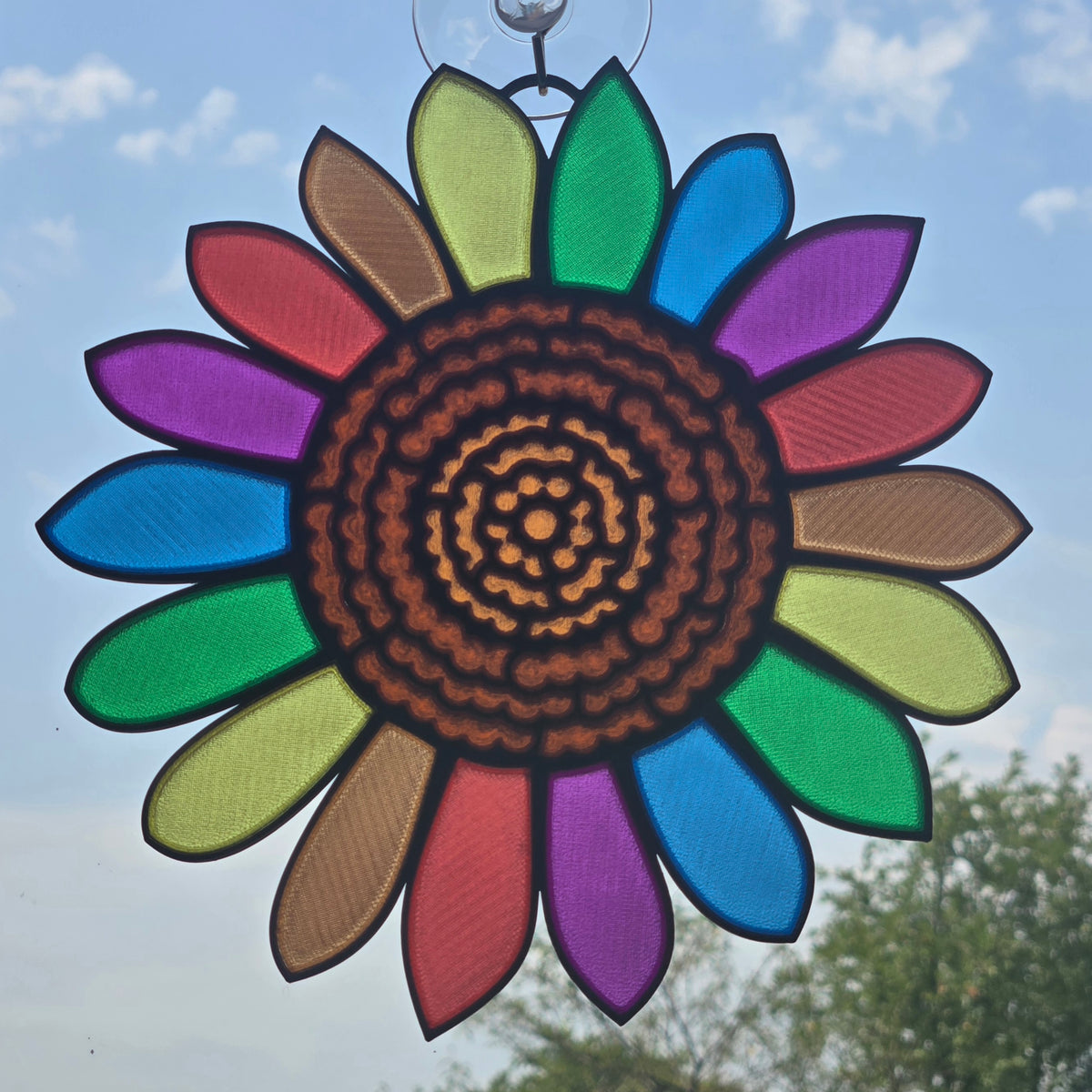 A colorful 3D printed sunflower suncatcher hanging in front of a blue sky with clouds and green trees in the background.