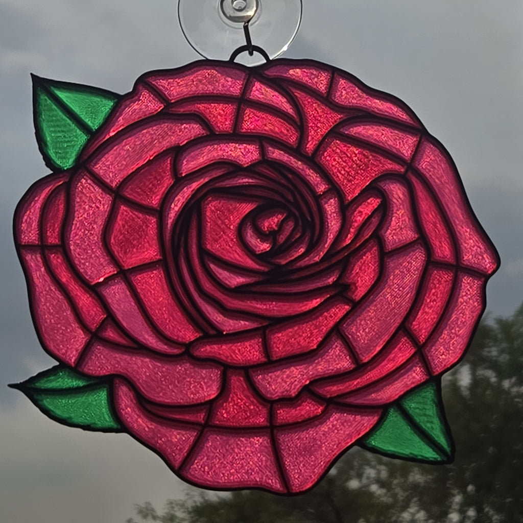 A 3D printed rose window ornament with vibrant pink and red glass petals, green leaves, and dark metal outlines, suspended by a clear hanging loop against an outdoor background.