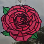 A 3D printed rose window ornament with vibrant pink and red glass petals, green leaves, and dark metal outlines, suspended by a clear hanging loop against an outdoor background.