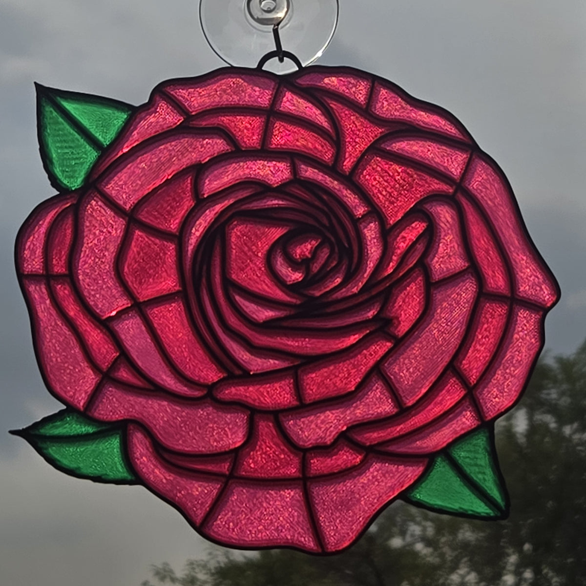 A 3D printed rose window ornament with vibrant pink and red glass petals, green leaves, and dark metal outlines, suspended by a clear hanging loop against an outdoor background.