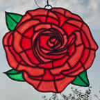 A 3D printed rose window hanging ornament featuring a red rose with green leaves, suspended by a metal loop against a cloudy sky.