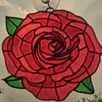 A red rose 3D printed suncatcher with green leaves and a clear suction cup at the top, shown hanging against an outdoor sky background.