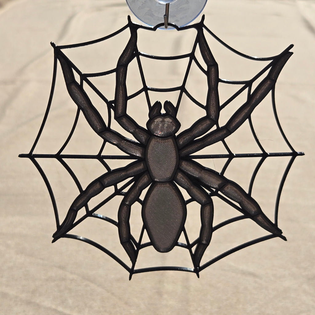 A black spider web hanging ornament with a detailed spider in the center, suspended from a clear hook at the top against a beige fabric background.