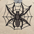 A black spider web hanging ornament with a detailed spider in the center, suspended from a clear hook at the top against a beige fabric background.