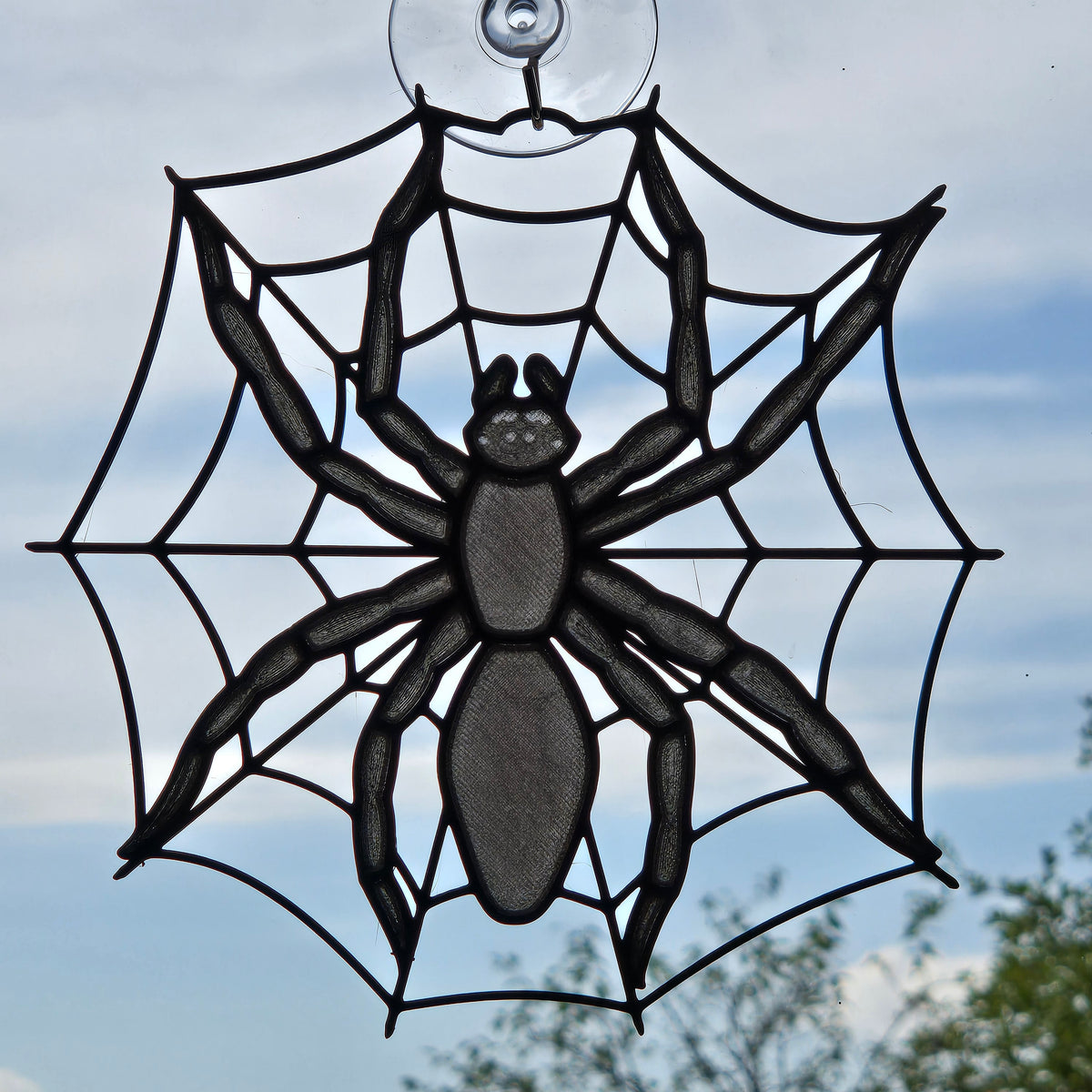 A black spider web suncatcher with a large spider in the center and a clear suction cup at the top, shown against an outdoor sky background.