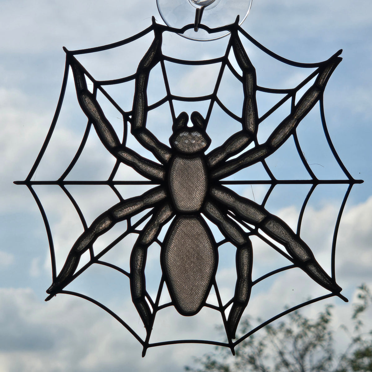 A black spider web suncatcher with a large spider in the center, shown hanging in front of a cloudy sky.