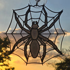 A black spider web suncatcher with a large spider silhouette, attached to a clear suction cup on a window, set against a warm sunset sky with trees in the background.