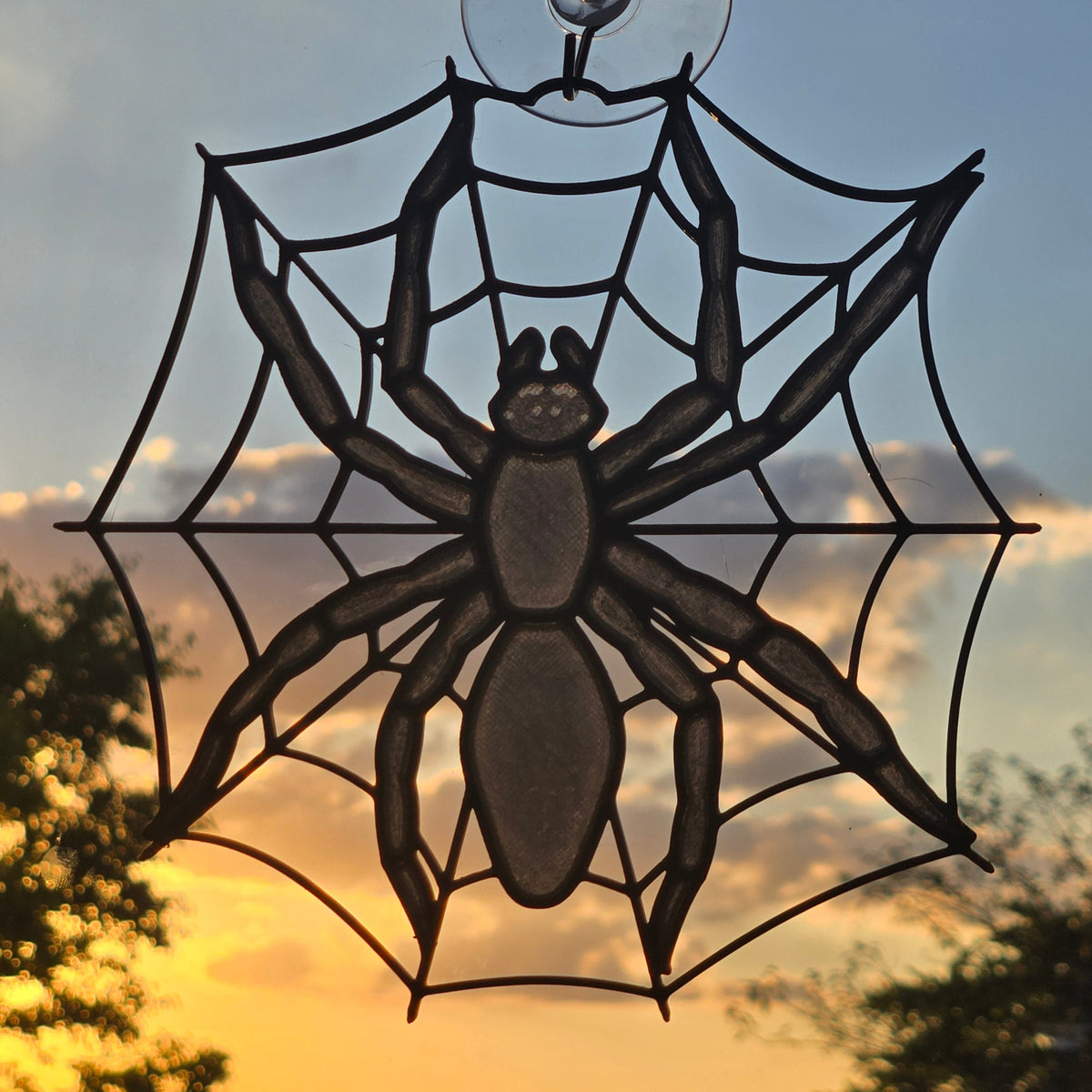 A black spider web suncatcher with a large spider silhouette, attached to a clear suction cup on a window, set against a warm sunset sky with trees in the background.