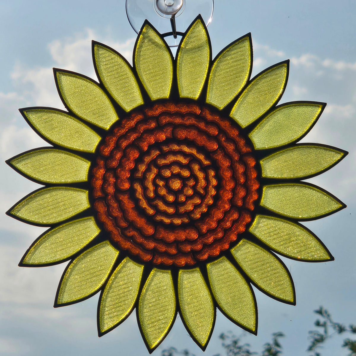 A 3D printed sunflower hanging ornament with a textured orange-brown center and yellow-green petals, suspended by a clear hook against a blue sky background.