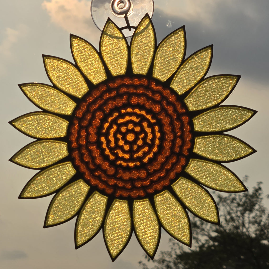 A #D printed sunflower hanging ornament with textured yellow petals and a dark brown patterned center, suspended by a clear loop against an outdoor sky background.