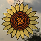 A #D printed sunflower hanging ornament with textured yellow petals and a dark brown patterned center, suspended by a clear loop against an outdoor sky background.