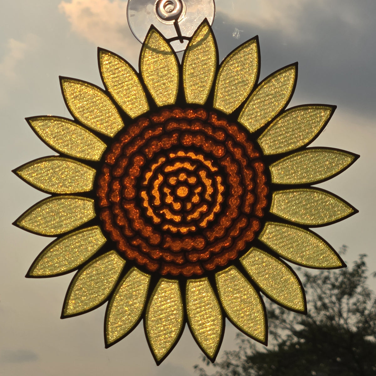 A #D printed sunflower hanging ornament with textured yellow petals and a dark brown patterned center, suspended by a clear loop against an outdoor sky background.